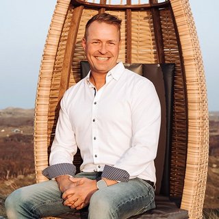 Ein lächelnder Mann sitzt entspannt in einem modernen Korbsessel, umgeben von einer malerischen Landschaft.