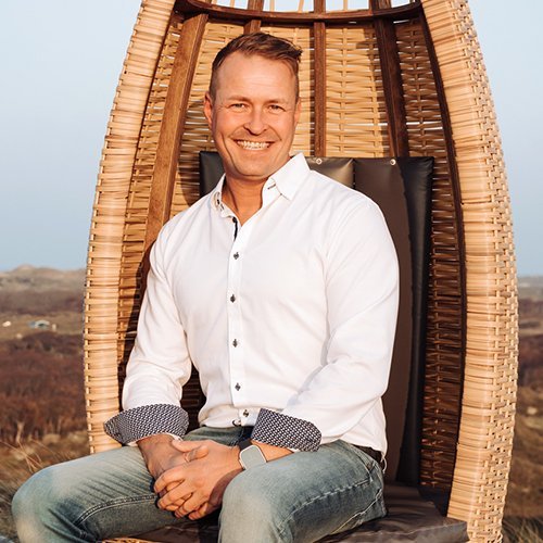 Ein lächelnder Mann sitzt entspannt in einem modernen Korbsessel, umgeben von einer malerischen Landschaft.