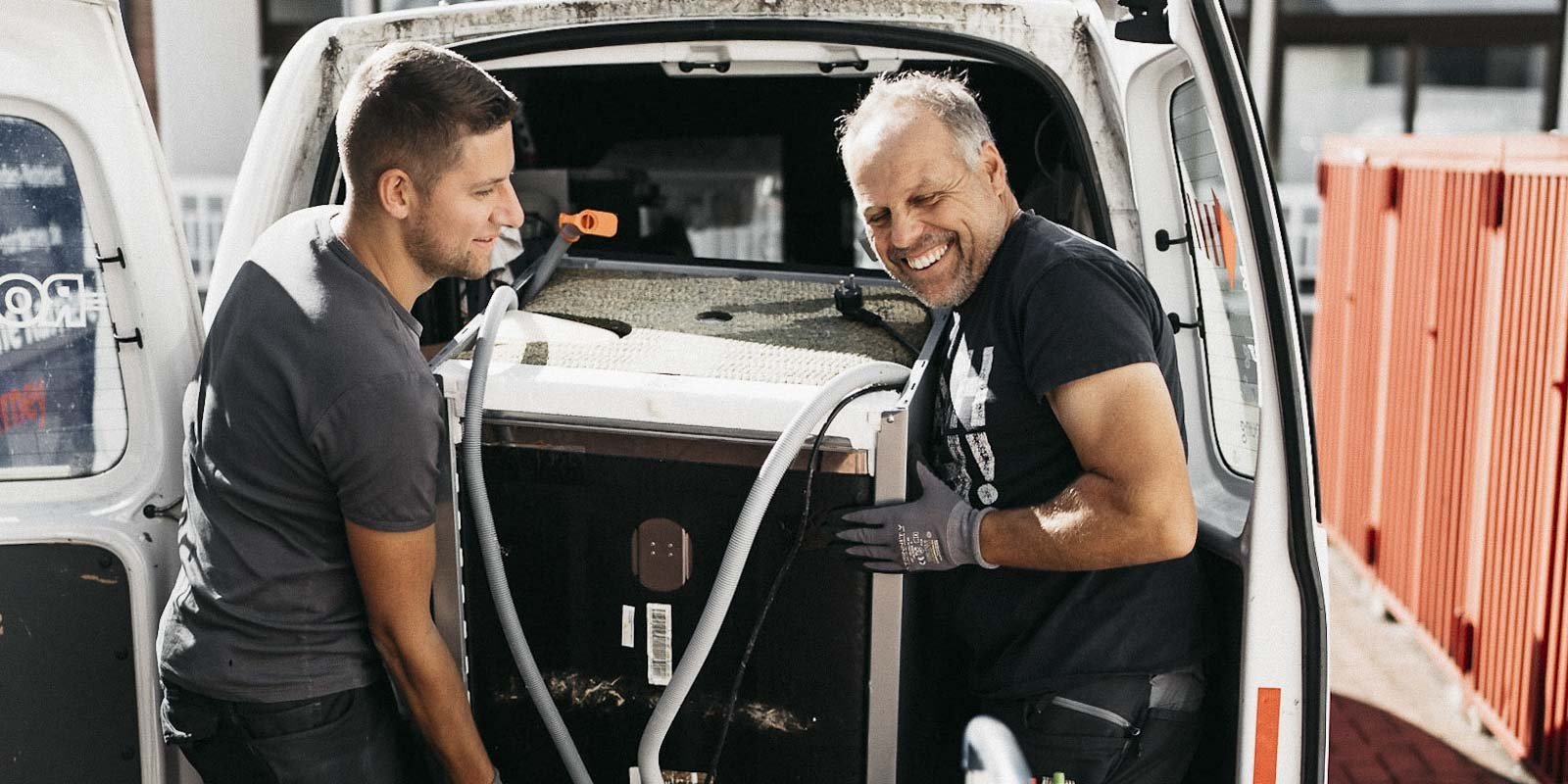 Zwei Männer heben gemeinsam ein großes Gerät aus einem Lieferwagen, beide lächeln und wirken zufrieden mit ihrer Arbeit.