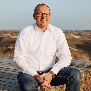 Ein Mann mit Brille sitzt entspannt auf einer Holzplattform und lächelt, während die Dünen im Hintergrund sichtbar sind.