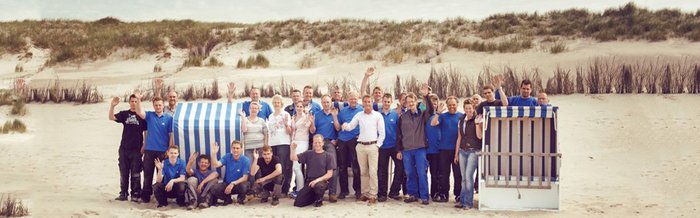 Eine Gruppe von Menschen in blauen T-Shirts winkt fröhlich am Strand, umgeben von Sand und Dünen.