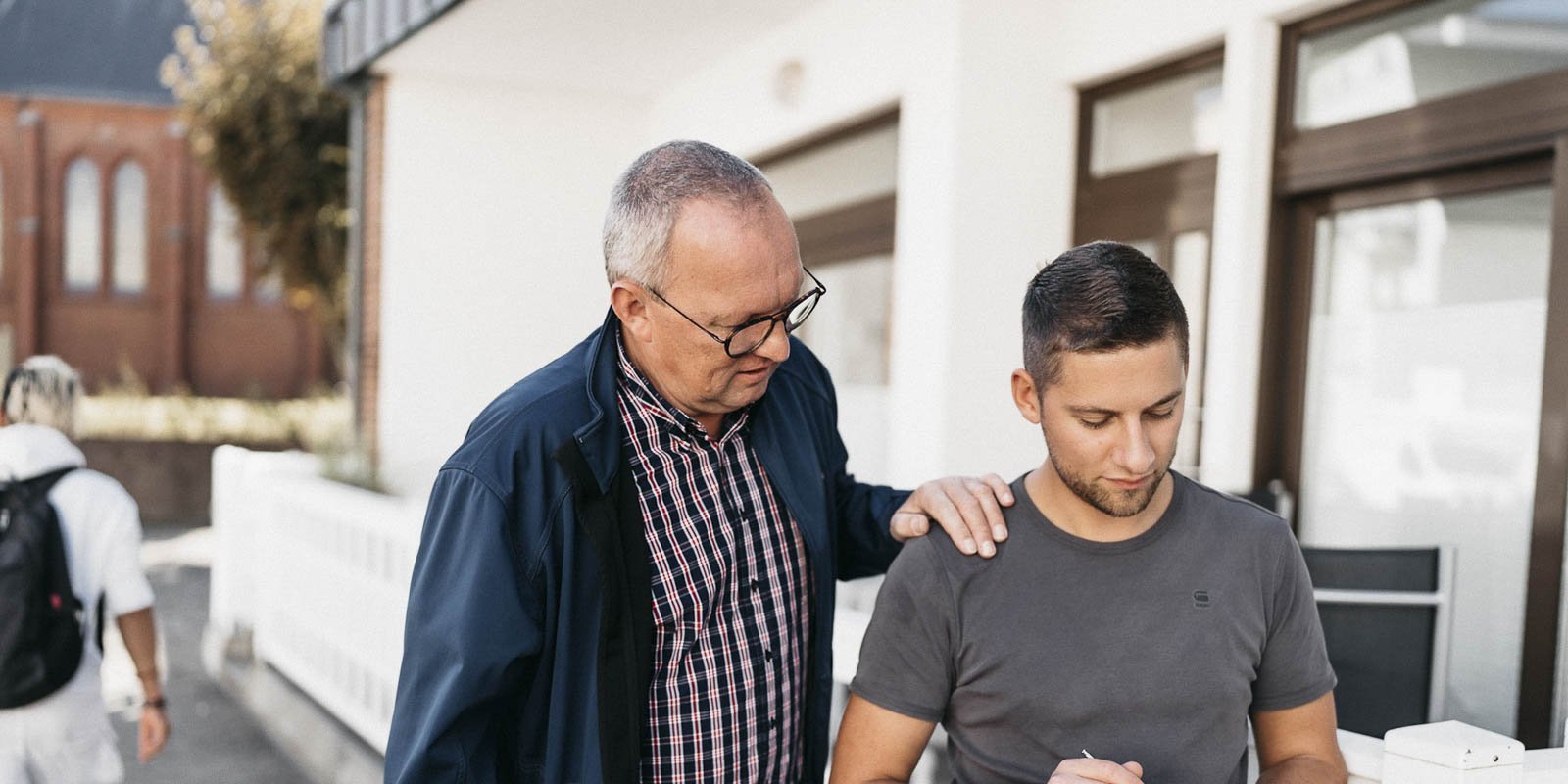 Ein älterer Mann steht neben einem jüngeren Mann, der konzentriert auf ein Blatt Papier schreibt, während er beobachtet wird.