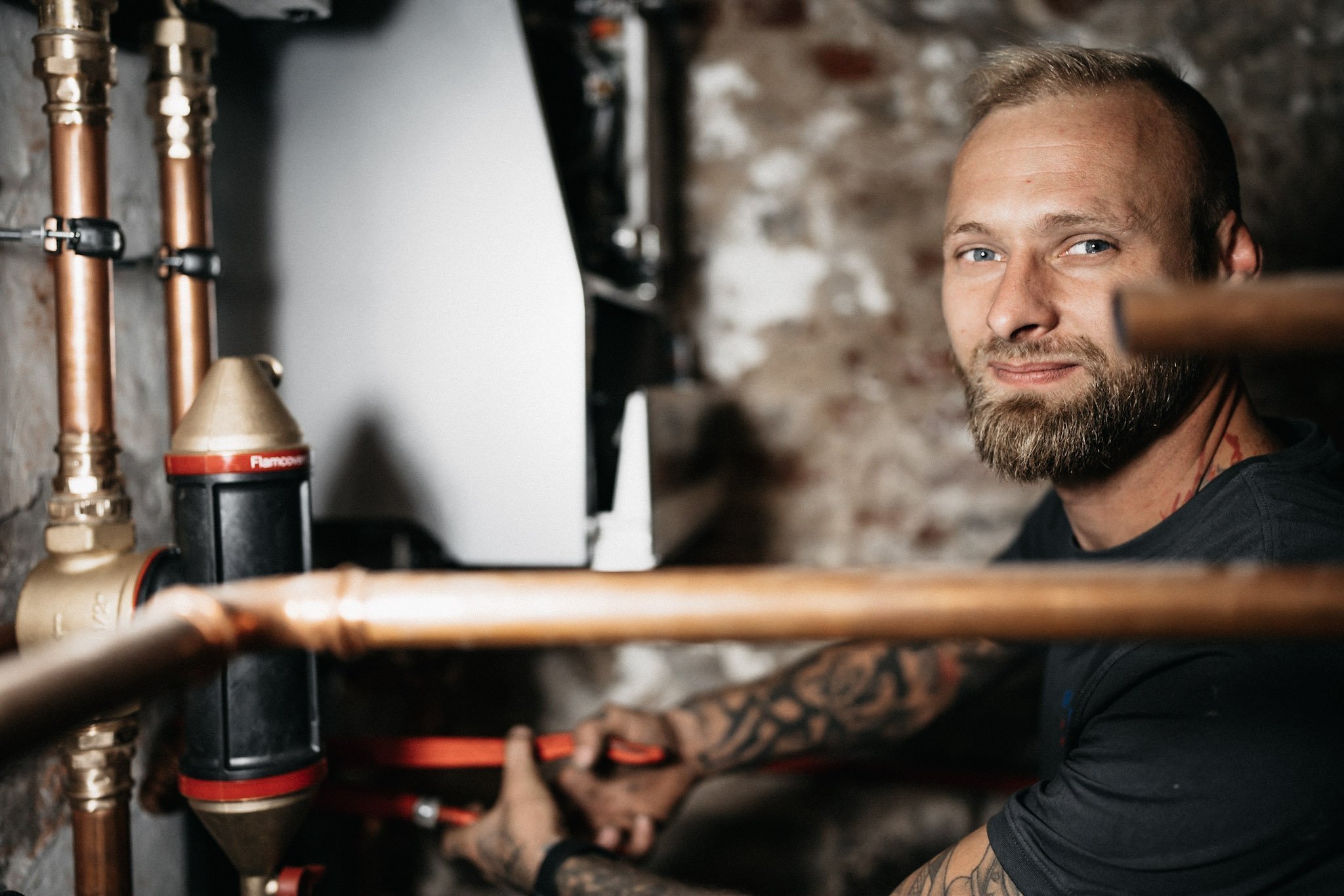 Ein Handwerker mit Bart arbeitet konzentriert an Rohren in einem Keller, umgeben von Kupferleitungen und einer steinernen Wand.
