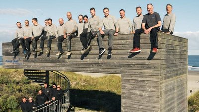 Männer in grauen Shirts posieren fröhlich auf einer erhöhten Holzstruktur, während der Ozean im Hintergrund sichtbar ist.