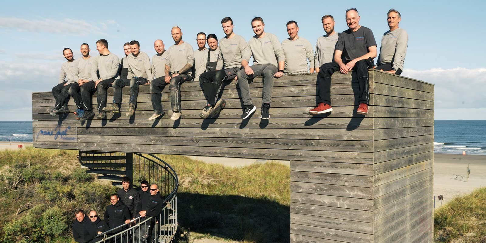 Männer in grauen Shirts posieren fröhlich auf einer erhöhten Holzstruktur, während der Ozean im Hintergrund sichtbar ist.