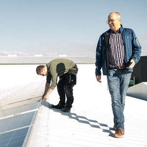 Auf dem Dach eines Gebäudes repariert ein junger Mann mit Werkzeug, während ein älterer Mann lächelnd zuschaut.