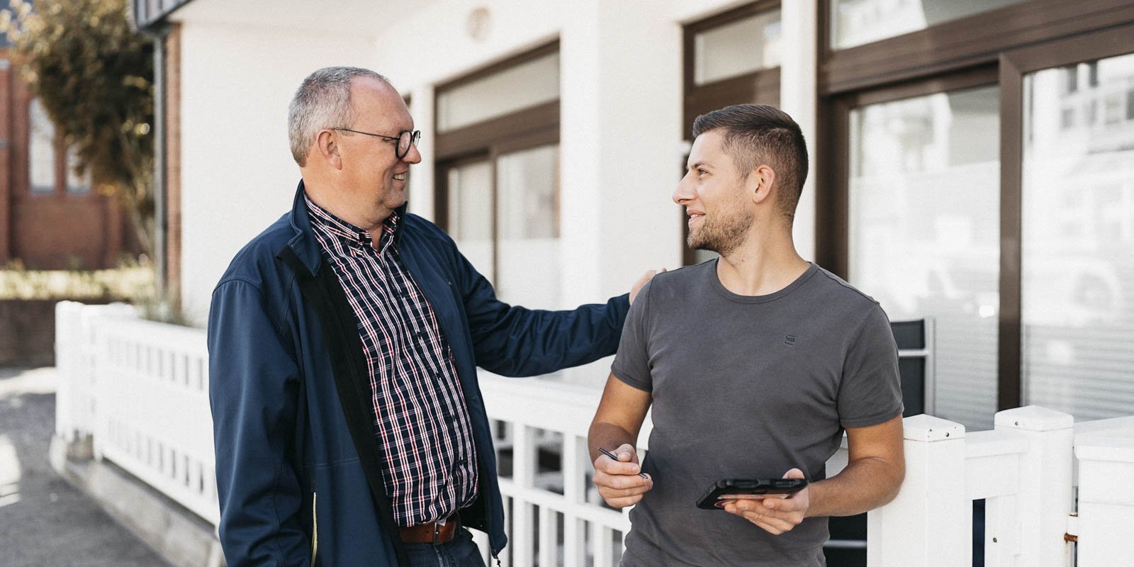 Ein älterer Mann spricht freundlich mit einem jüngeren Mann, während sie an einem hellen Gebäude stehen.