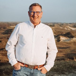 Ein lächelnder Mann in einem weißen Hemd steht in einer hügeligen Landschaft mit sanften Erhebungen im Hintergrund.