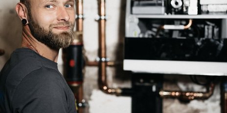 In einem technischen Raum lächelt ein junger Handwerker, während er an einer modernen Heizungsanlage arbeitet.
