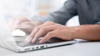 Eine Person tippt konzentriert auf einem Laptop, während ein Cloud-Symbol im Hintergrund sichtbar ist.