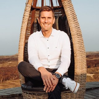 In einer malerischen Umgebung sitzt ein lächelnder Mann in einem modernen Korbstuhl, umgeben von sanften Hügeln.