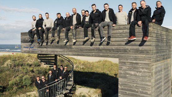 Männer in Arbeitskleidung posieren auf einer erhöhten Holzstruktur, umgeben von Dünen und einem klaren Himmel.