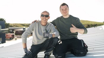 Auf einem sonnigen Dach sitzen zwei Männer in Arbeitskleidung, beide strahlen Freude aus und zeigen positive Gesten.