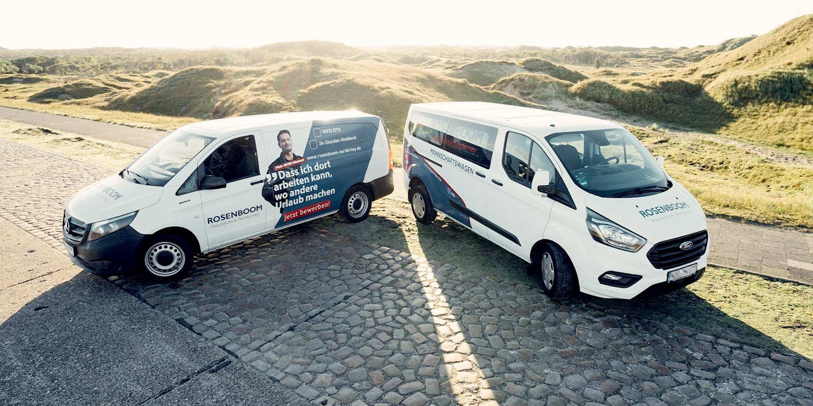 Zwei Lieferwagen stehen auf einem gepflasterten Weg, umgeben von sanften Hügeln und grünem Gras im Hintergrund.