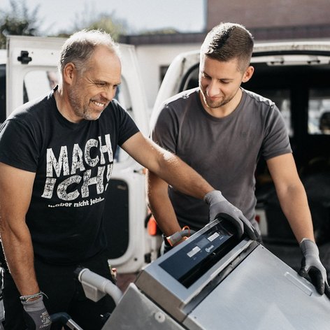 Zwei Männer lächeln, während sie gemeinsam ein großes Gerät aus einem Lieferwagen heben, umgeben von Tageslicht.