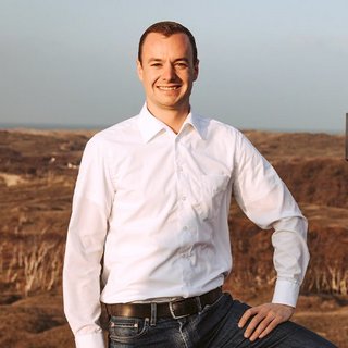 Ein junger Mann in einem weißen Hemd steht lächelnd in einer natürlichen Umgebung mit sanften Hügeln im Hintergrund.