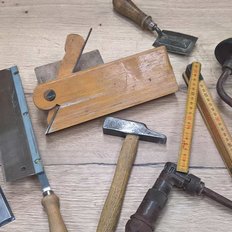 Auf einem Tisch sind verschiedene Handwerkzeuge angeordnet, darunter ein Meißel, eine Säge und ein Holzmaßstab.