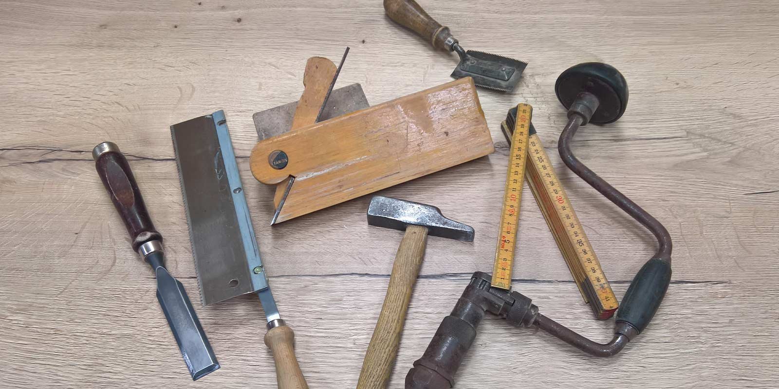 Auf einem Tisch sind verschiedene Handwerkzeuge angeordnet, darunter ein Meißel, eine Säge und ein Holzmaßstab.