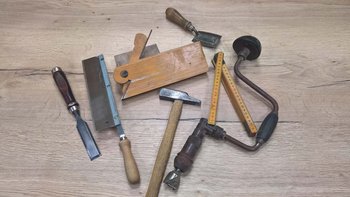 Eine Sammlung von Holzwerkzeugen liegt auf einem hellen Holztisch, darunter ein Hobel, ein Hammer und ein Maßstab.