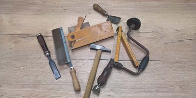 Eine Sammlung von Holzwerkzeugen liegt auf einem hellen Holztisch, darunter ein Hobel, ein Hammer und ein Maßstab.